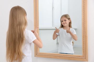 Photo of Beautiful little girl showing thumbs up while looking in mirror indoors