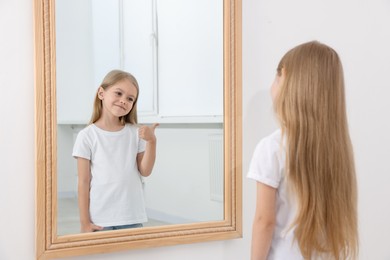 Photo of Beautiful little girl showing thumbs up while looking in mirror indoors