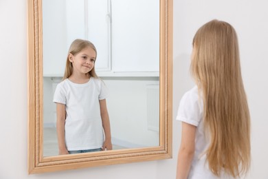 Photo of Beautiful little girl looking at herself in mirror indoors