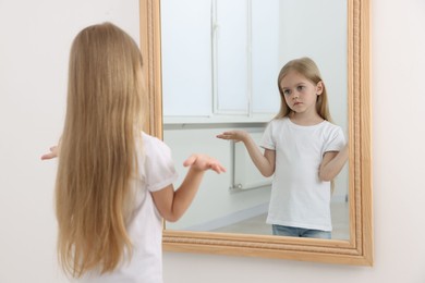 Photo of Upset little girl looking at herself in mirror indoors