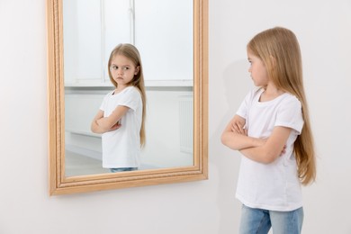 Photo of Upset little girl looking at herself in mirror indoors