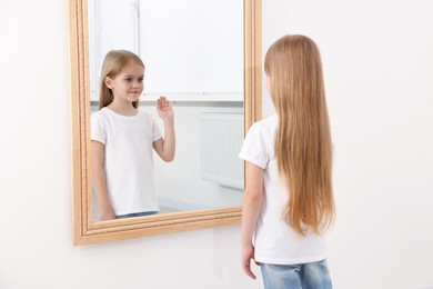 Photo of Beautiful little girl looking at herself in mirror indoors