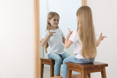 Photo of Beautiful little girl looking at herself in mirror indoors