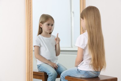 Photo of Upset little girl looking at herself in mirror indoors