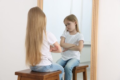 Photo of Upset little girl looking at herself in mirror indoors