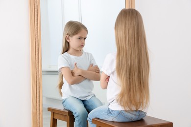Photo of Upset little girl looking at herself in mirror indoors