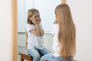 Photo of Beautiful little girl looking at herself in mirror indoors
