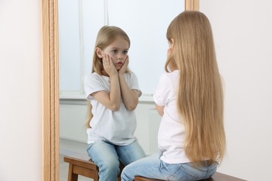 Photo of Upset little girl looking at herself in mirror indoors