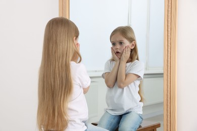 Photo of Upset little girl looking at herself in mirror indoors