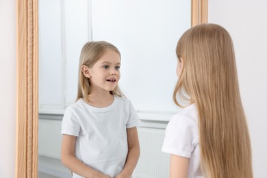 Photo of Beautiful little girl looking at herself in mirror indoors