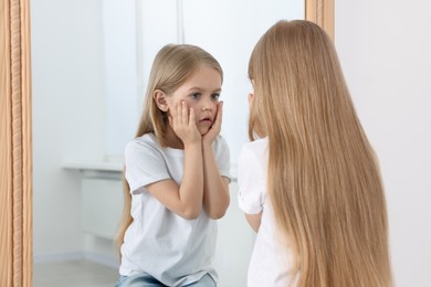 Photo of Upset little girl looking at herself in mirror indoors