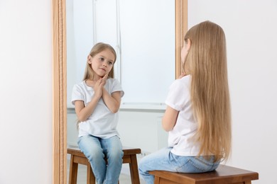 Photo of Upset little girl looking at herself in mirror indoors