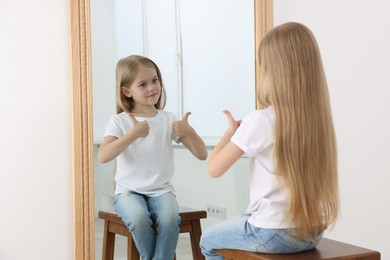Photo of Beautiful little girl showing thumbs up while looking in mirror indoors