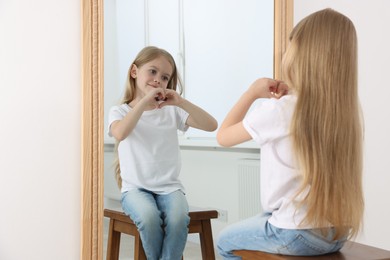 Photo of Beautiful little girl showing heart with hands while looking in mirror indoors