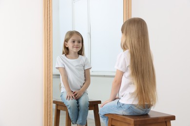 Photo of Beautiful little girl looking at herself in mirror indoors