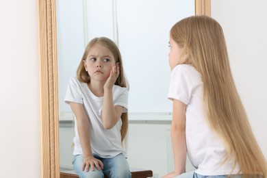 Photo of Upset little girl looking at herself in mirror indoors