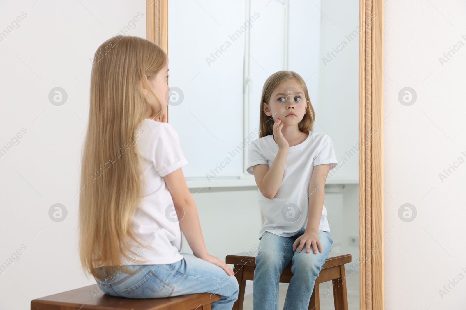 Upset little girl looking at herself in mirror indoors Photo of Upset little girl looking at herself in mirror indoors