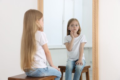 Photo of Upset little girl looking at herself in mirror indoors
