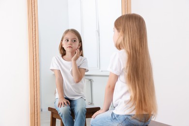 Photo of Upset little girl looking at herself in mirror indoors