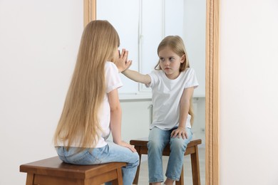 Photo of Upset little girl looking at herself in mirror indoors