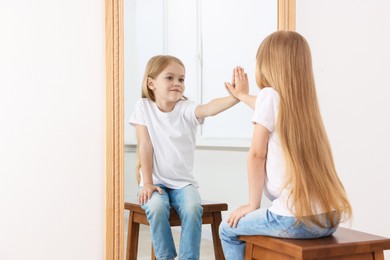 Photo of Beautiful little girl looking at herself in mirror indoors