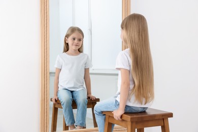 Photo of Beautiful little girl looking at herself in mirror indoors