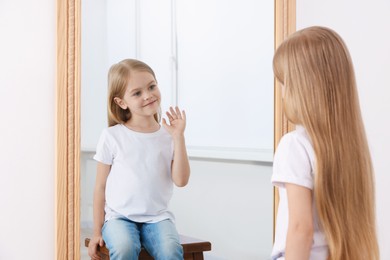 Photo of Beautiful little girl waving while looking in mirror indoors