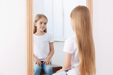 Photo of Upset little girl looking at herself in mirror indoors