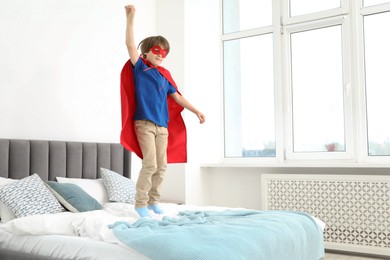 Photo of Cute little boy in superhero costume jumping on bed at home