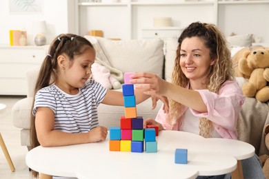 Photo of Mother and her daughter building tower with colorful cubes at white table indoors