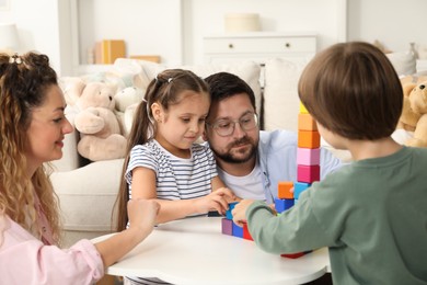 Photo of Children and their parents building tower with colorful cubes at white table indoors