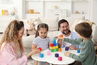 Photo of Children and their parents building tower with colorful cubes at white table indoors