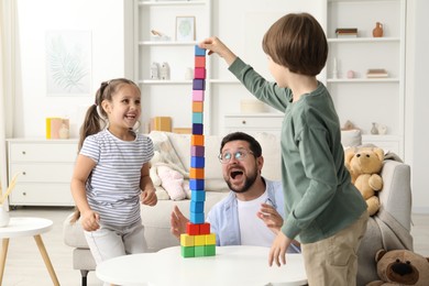 Photo of Children and their father building tower with colorful cubes at white table indoors