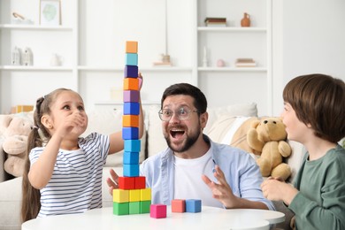 Photo of Children and their father building tower with colorful cubes at white table indoors