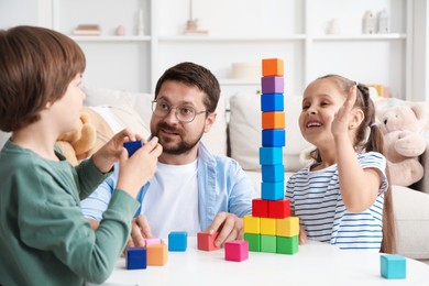 Photo of Children and their father building tower with colorful cubes at white table indoors