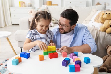 Photo of Father and his daughter playing with colorful cubes at white table indoors