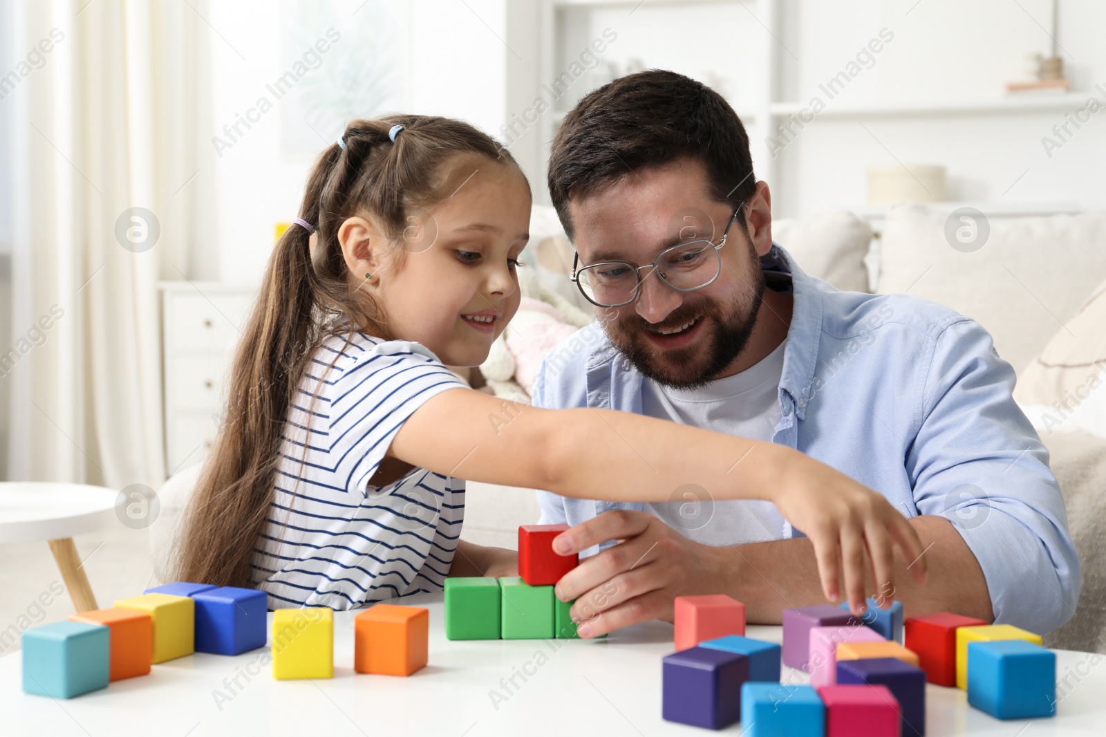 Father and his daughter playing with colorful cubes at white table indoors Photo of Father and his daughter playing with colorful cubes at white table indoors