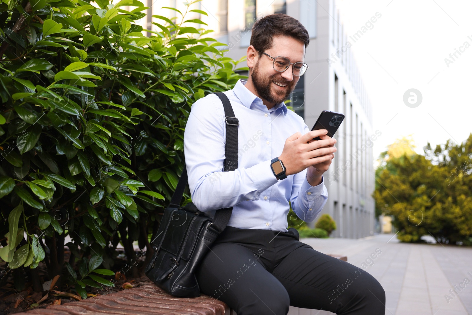 Successful businessman with smartphone on bench near office building Photo of Successful businessman with smartphone on bench near office building