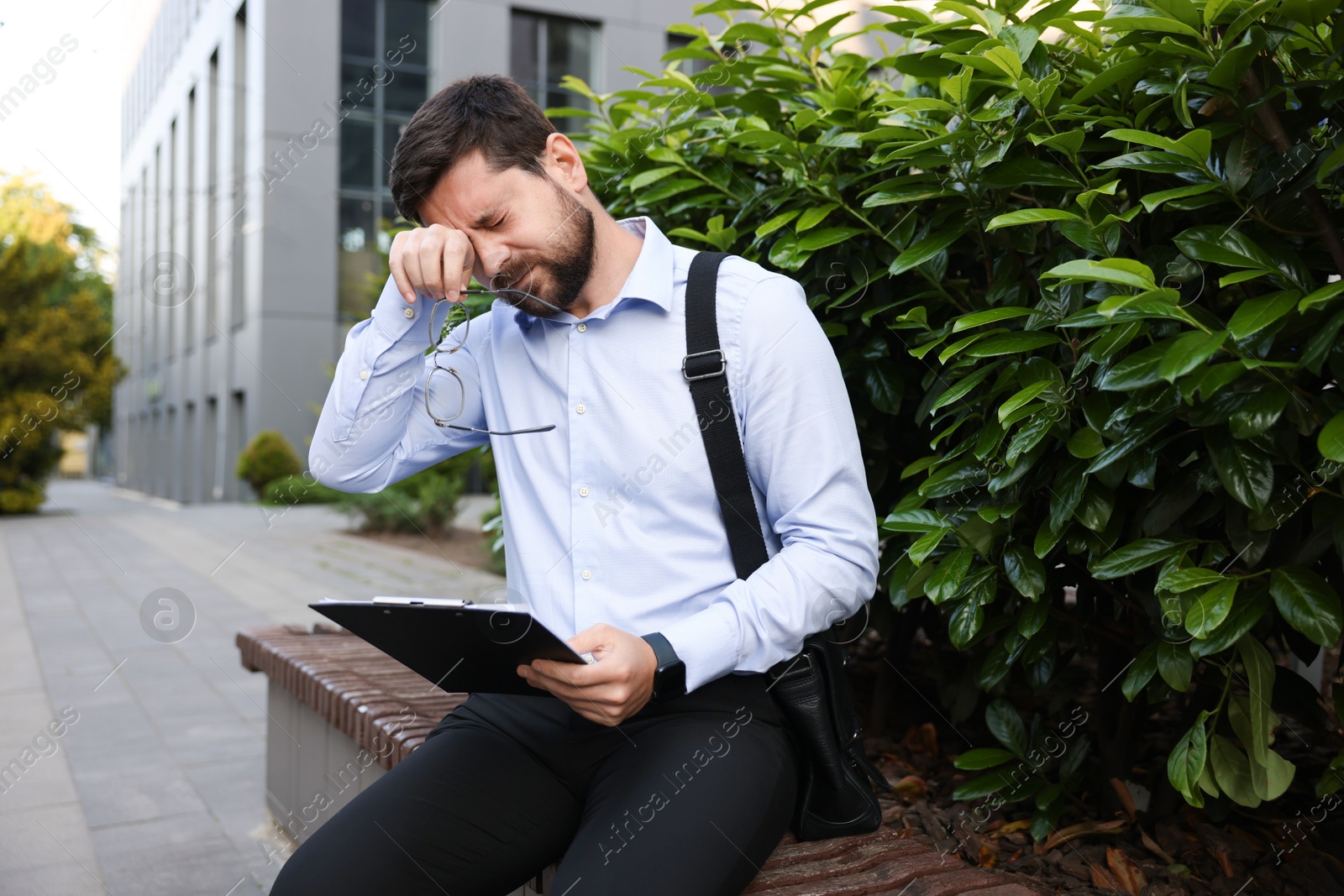 Tired businessman with glasses and clipboard outside office building Photo of Tired businessman with glasses and clipboard outside office building