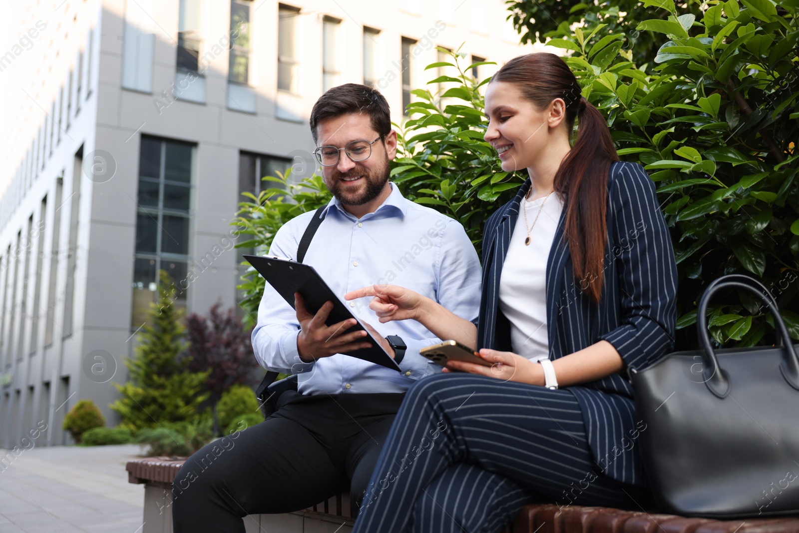 Happy coworkers discussing something on bench near office building Photo of Happy coworkers discussing something on bench near office building