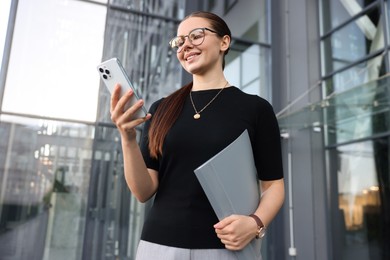 Photo of Successful businesswoman with smartphone and folder near office building, low angle view