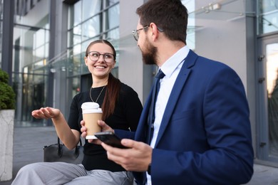 Photo of Happy coworkers with smartphone and coffee discussing something near office building