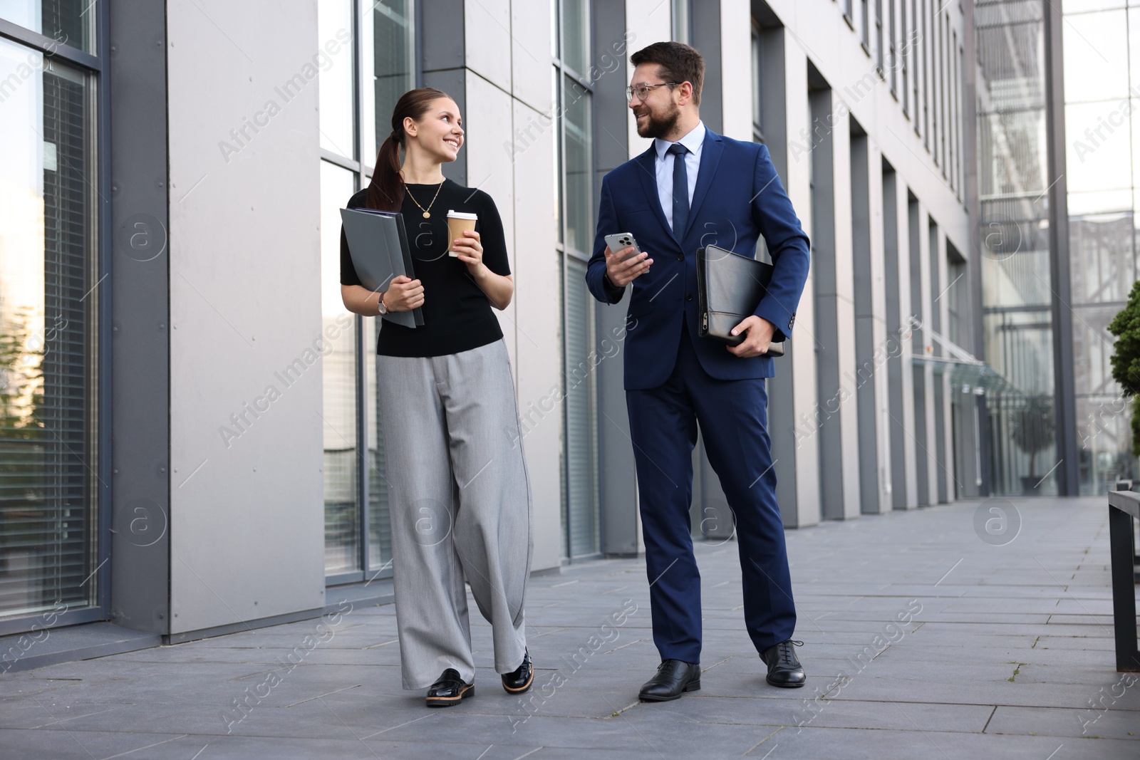 Happy coworkers with smartphone and coffee discussing something near office building Photo of Happy coworkers with smartphone and coffee discussing something near office building