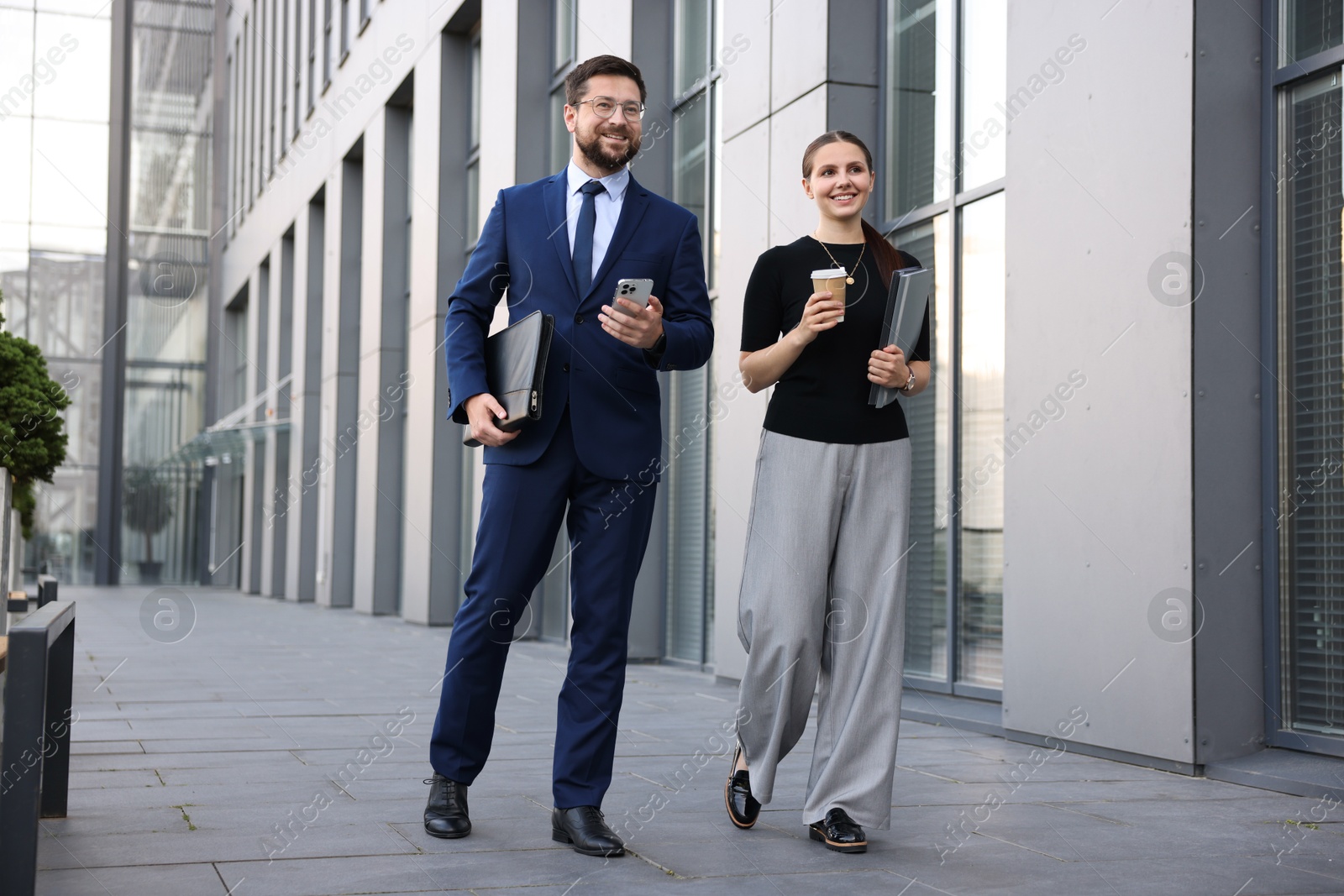 Happy coworkers with smartphone and coffee discussing something near office building Photo of Happy coworkers with smartphone and coffee discussing something near office building