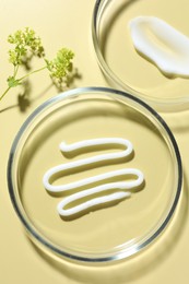 Photo of Different cosmetic products in Petri dishes and floral decor on pale yellow background, flat lay