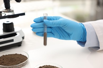 Photo of Scientist with test tube of soil sample at white table in laboratory, closeup