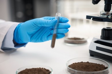 Photo of Scientist with test tube of soil sample at white table in laboratory, closeup