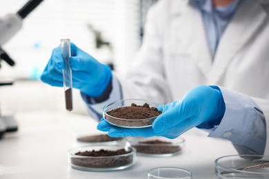 Photo of Scientist holding Petri dish with soil sample and test tube at white table in laboratory, closeup