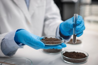 Photo of Scientist holding Petri dish with soil sample and test tube at white table in laboratory, closeup