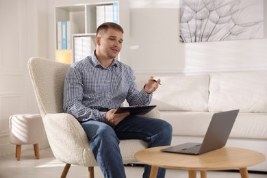 Photo of Smiling psychologist with clipboard during online session by laptop in office